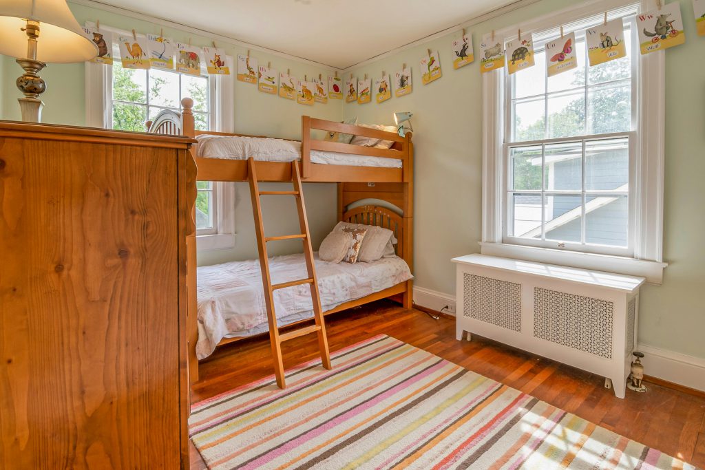 Zdjęcie dwupiętrowego łóżka z białą pościelą w jasnym pokoju dziecięcym; łóżko wykonane z drewna, z solidną drabinką i zabezpieczeniami na górnym poziomie, otoczone zabawkami i kolorowymi dekoracjami.